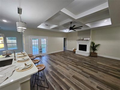 Living room with ceiling fan, dark luxury vinyl looking wood-type flooring, tray box beam ceiling, a wood brick fireplace, and view of kitchen island looking out the pool.