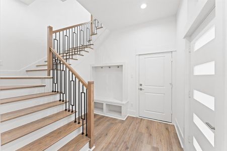 This photo shows a modern entryway with a wooden staircase featuring sleek black metal railing. The space is bright and airy, with light wood flooring and white walls. There's a built-in bench with hooks for storage and a white door with horizontal glass panels.