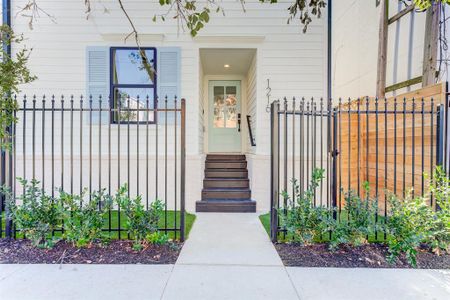 This photo showcases a modern fenced Victorian home entrance with a stylish door, black accented double paned windows, adjacent to a matching garage door. The exterior features clean white Hardie Plank siding neutral brick, and a small landscaped area with shrubs.