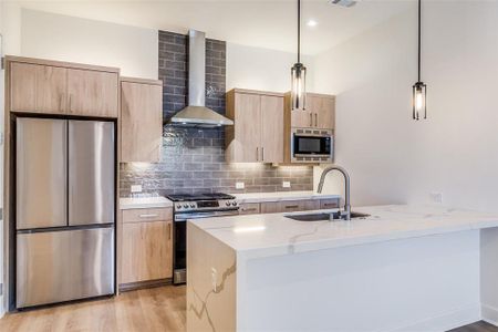 Kitchen with wall chimney range hood, light hardwood / wood-style flooring, stainless steel appliances, light stone counters, and decorative light fixtures