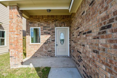 A brick house entryway featuring a white door with oval glass, a small window, and a concrete porch under a shaded overhang.