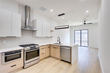 Kitchen with light wood-type flooring, stainless steel appliances, wall chimney exhaust hood, kitchen peninsula, and light brown cabinetry