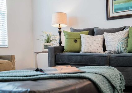 This living room is decorated with a dark gray sofa with green and light colored pillows, throw blanket with magazine on it, and framed painting on the wall.