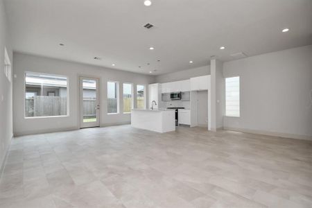 This is a spacious, modern open-plan living area with a bright kitchen, featuring large windows for ample natural light. The room offers direct access to the backyard and is finished with light tile flooring throughout.