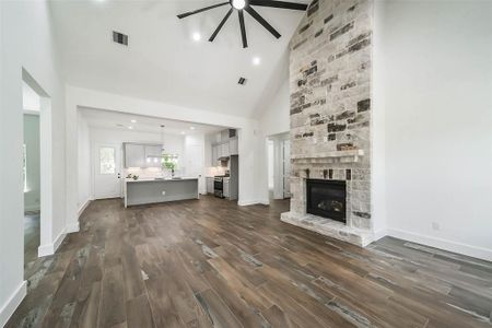 This modern open-concept living space features a high vaulted ceiling with a striking fan, a stone fireplace as a focal point, and a spacious kitchen with an island and stainless steel appliances. The hardwood floors add warmth and elegance.