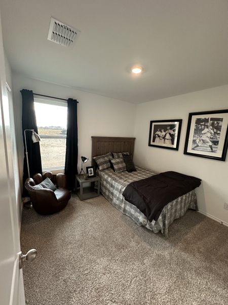 A cozy bedroom with plaid bedding, framed artwork, and a comfy leather chair. Neutral carpet and natural light complete the scene.