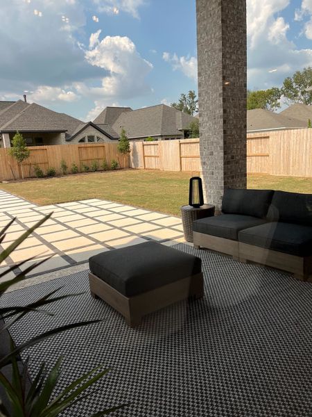 A serene patio with modern outdoor furniture, patterned rug, and spacious backyard under a partly cloudy sky.