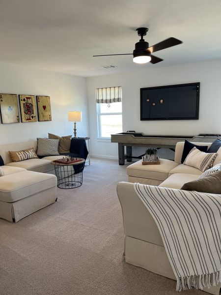 A cozy living room with beige seating, modern fan, game table, and playful wall art. Perfect for relaxing and entertaining. A cozy living room with beige seating, modern fan, game table, and playful wall art. Perfect for relaxing and entertaining.