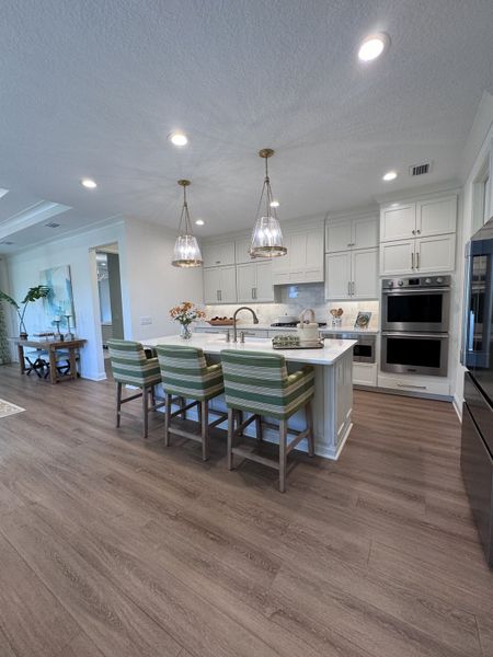 A modern kitchen featuring a spacious island, pendant lights, sleek cabinetry, and striped bar stools on wooden flooring.
