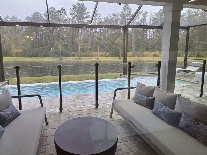 A cozy patio with comfortable seating, overlooking a serene pool and river view, enclosed in a glass-paneled area.