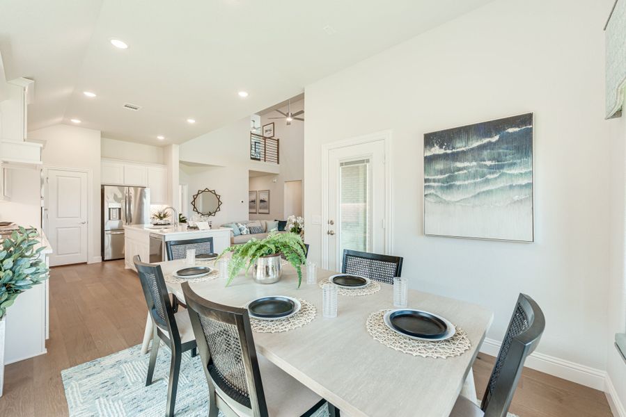 Open dining area with white table and four chairs, hardwood floors, and kitchen visible in background Open dining area with white table and four chairs, hardwood floors, and kitchen visible in background