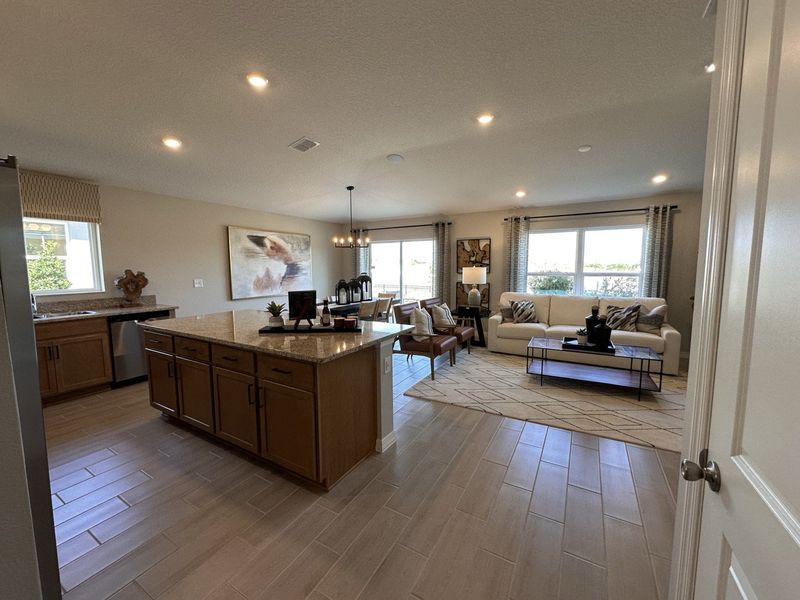 A spacious open-concept kitchen and living area featuring wood cabinetry, granite countertops, and warm-toned flooring in Landings at Lake Mabel Loop by KB Home (Dundee, FL).