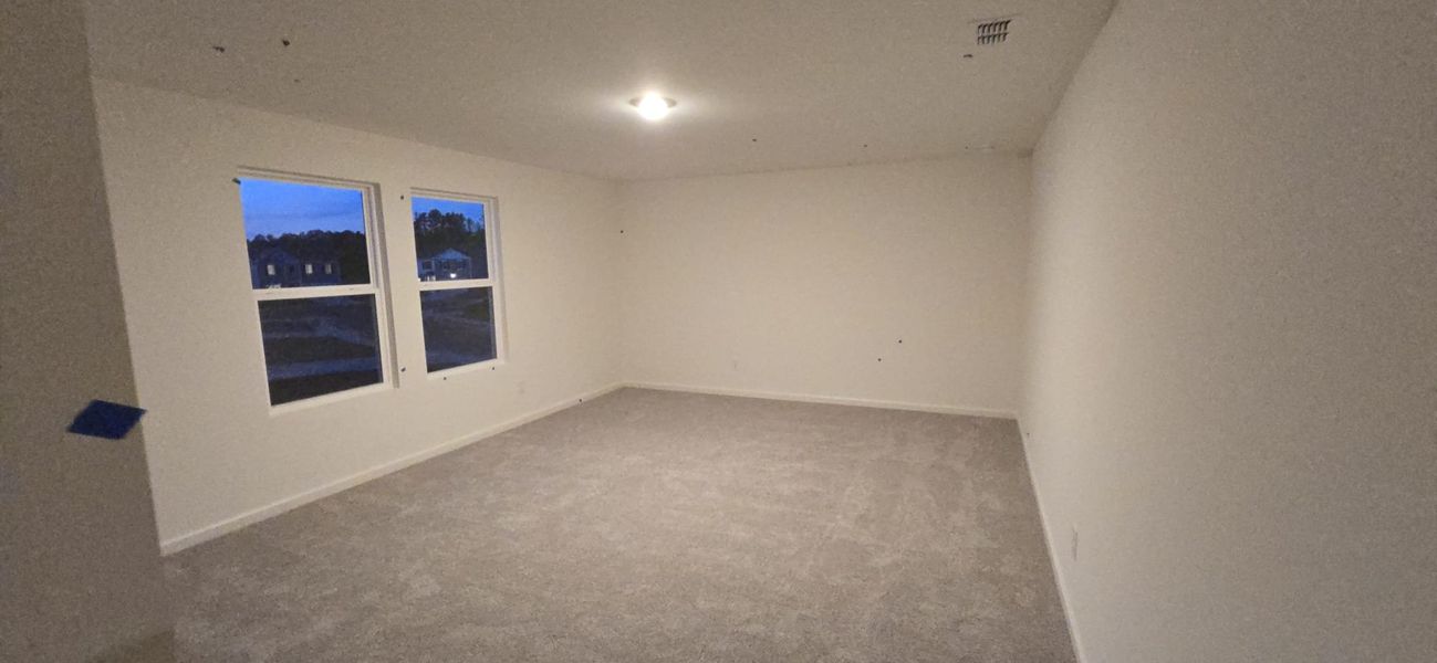 A spacious room with soft carpeting, neutral walls, and large windows. Cozy lighting enhances its inviting feel.