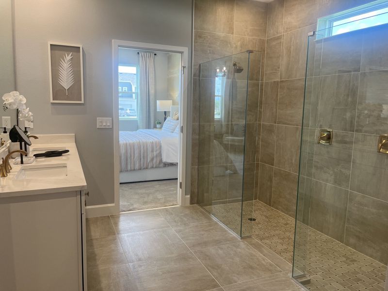 A modern bathroom features a glass shower, large tiles, and a sleek vanity, leading into a bright, cozy bedroom.
