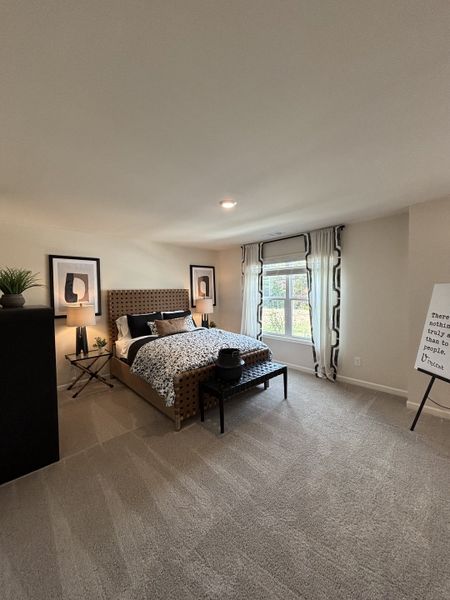 A cozy bedroom featuring a patterned bedspread, elegant artwork, and soft carpeting with natural light from a large window.