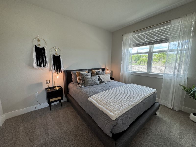 A cozy bedroom with a plush gray bed, elegant lighting, and large window with sheer curtains.