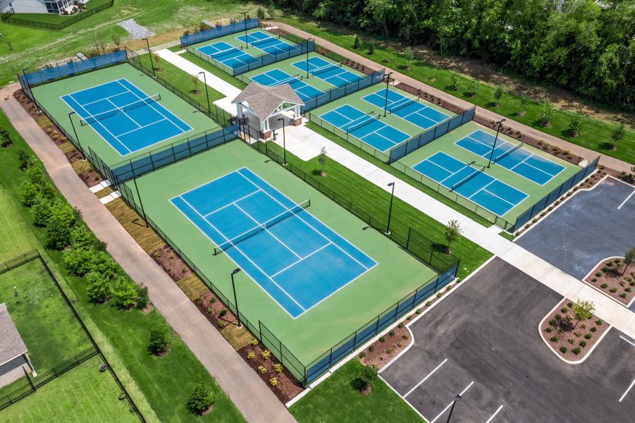 Aerial view of six tennis courts with green and blue surfaces, fencing, a small pavilion, and adjacent parking lot.