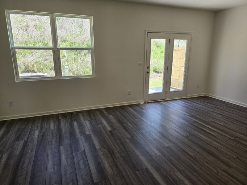 A bright living area with dark wood flooring, large windows, and French doors opening to a lush backyard.