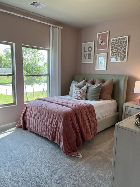 A cozy bedroom with pastel walls, soft carpeting, a plush bed with pink accents, and framed wall art.