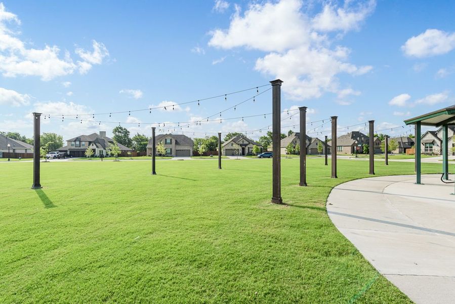 Community green space with string lights on poles, curved walkway, and neighborhood homes in background Community green space with string lights on poles, curved walkway, and neighborhood homes in background