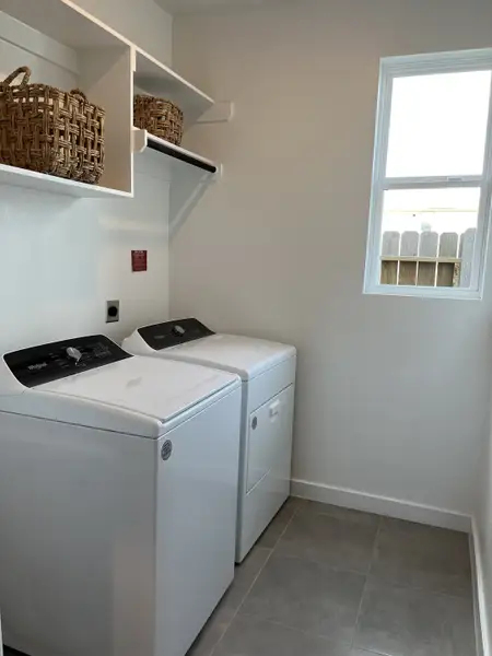 A functional laundry room with a washer, dryer, open shelving, and a bright window.
