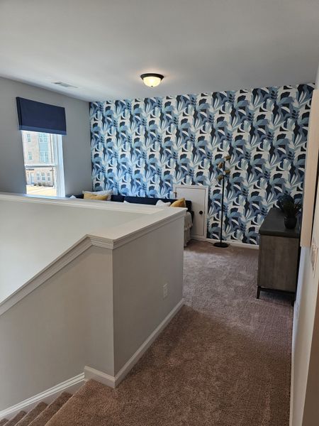 A cozy loft area with floral accent wallpaper, plush carpet, and natural light through a large window.