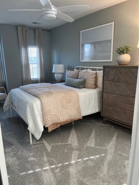 A cozy bedroom with plush carpeting, elegant bedding, a modern ceiling fan, and stylish decor accents.