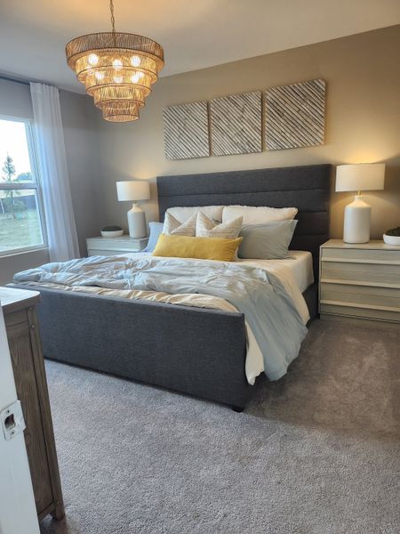 A cozy bedroom with a plush gray bed, chic chandelier, and modern decor accents in neutral tones.