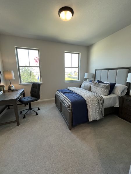 A cozy bedroom with a plush bed, elegant desk, and soft carpeting, bathed in natural light from two large windows.