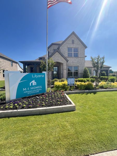 A beautiful brick home with lush landscaping in Lilyana by M/I Homes (Prosper, TX), highlighted by a vibrant green lawn.