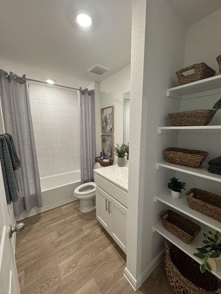 A cozy bathroom with a white vanity, tile bathtub, wood floors, and wicker baskets on open shelves. A cozy bathroom with a white vanity, tile bathtub, wood floors, and wicker baskets on open shelves.