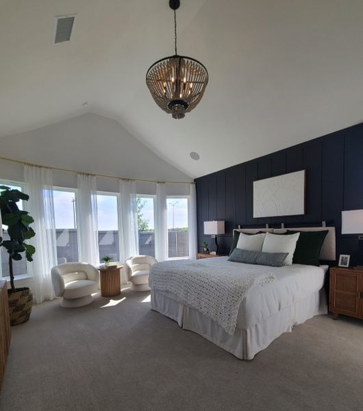 A spacious bedroom with a vaulted ceiling, dark accent wall, modern chandelier, and cozy seating by large bay windows.