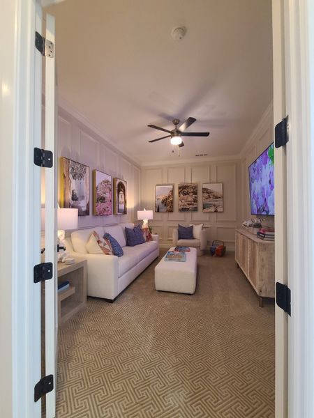 A cozy living room with a plush white sofa, textured carpet, ceiling fan, and artistic wall decor.