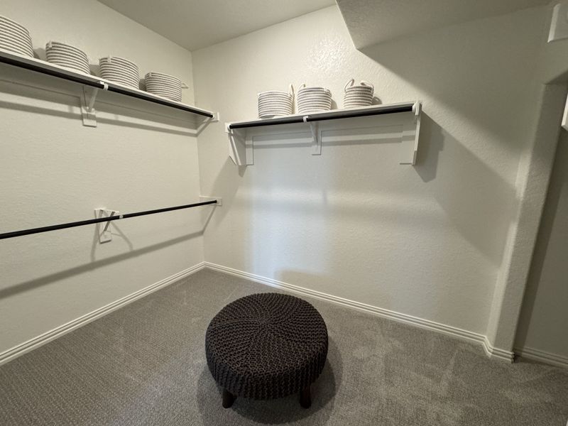 A spacious walk-in closet featuring carpet flooring, organized shelving, and a cozy textured ottoman for seating. A spacious walk-in closet featuring carpet flooring, organized shelving, and a cozy textured ottoman for seating.