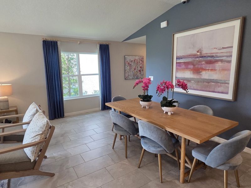 A cozy dining area with a wooden table, plush chairs, vibrant artwork, and bright windows.