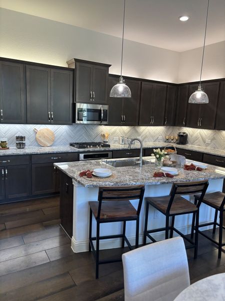 A modern kitchen with dark cabinets, granite countertops, a tiled backsplash, and sleek pendant lighting.
