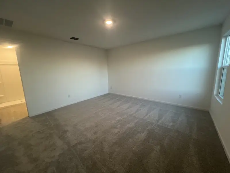 A spacious bedroom with soft carpeting, neutral walls, and ample natural light from a single window.