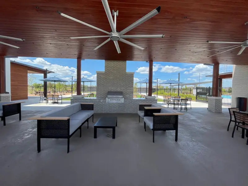 A modern outdoor lounge with cozy seating, fans, and a grill area under a wooden ceiling, near a pool.