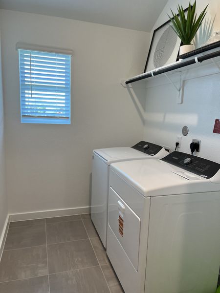 A modern laundry room with sleek appliances, gray tile flooring, a shelf, and a window for natural light.