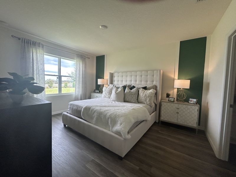 A cozy bedroom with a tufted bed, green accent walls, soft lighting, and large windows overlooking a serene view.