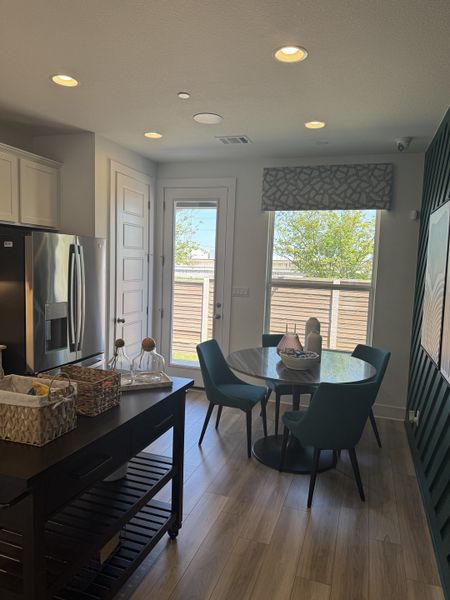 A modern dining area with a round glass table, teal chairs, and a sleek kitchen cart under recessed lighting.