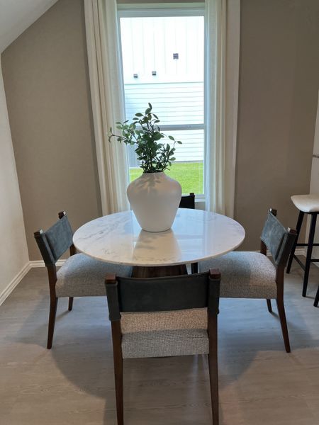 A cozy dining nook with a round marble table, elegant chairs, and a large vase by a bright window.