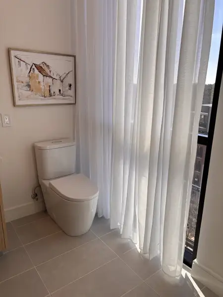 A modern bathroom with sleek tile flooring, a contemporary toilet, and soft sheer curtains.