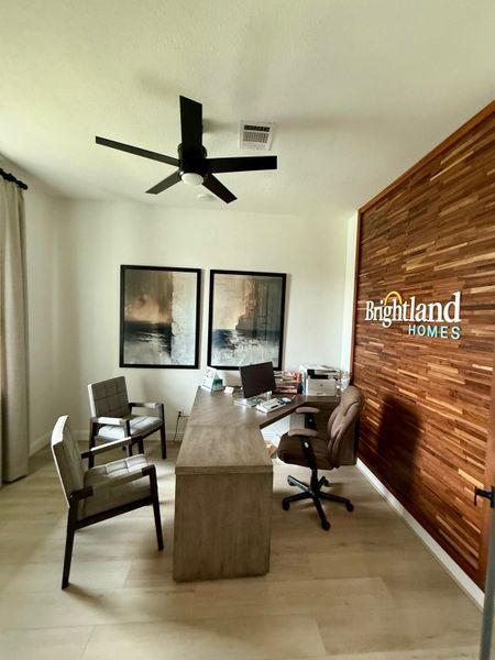 A modern office with a sleek wooden wall, minimalist desk, and cozy seating, accented by contemporary art and a ceiling fan.