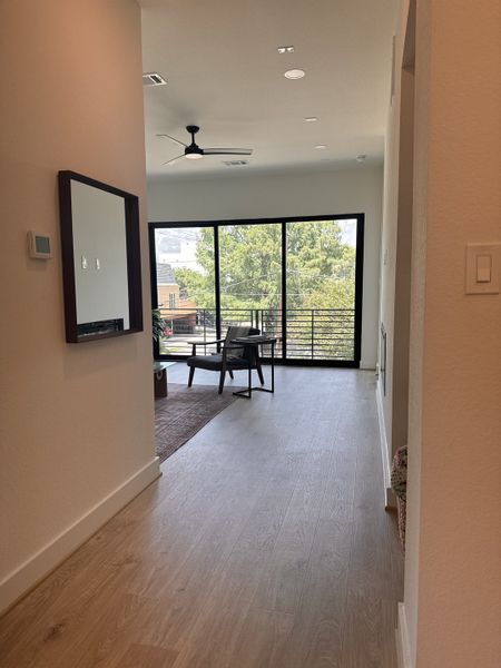 A modern hallway with wooden floors leading to a bright living area featuring large windows and a sleek ceiling fan.