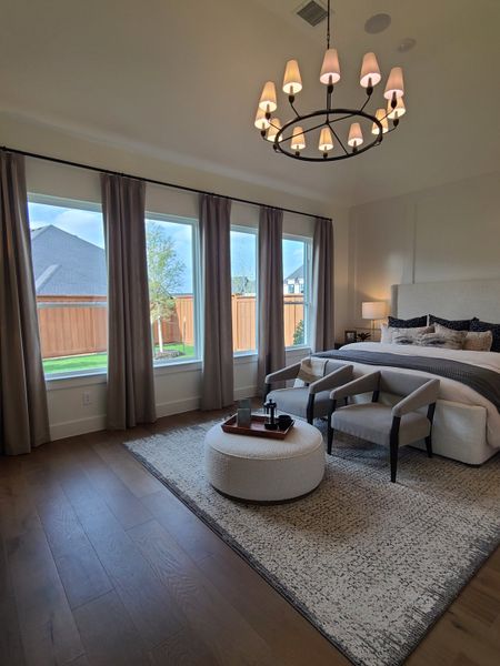 A cozy bedroom with elegant chandeliers, large windows, plush seating, and a textured area rug.
