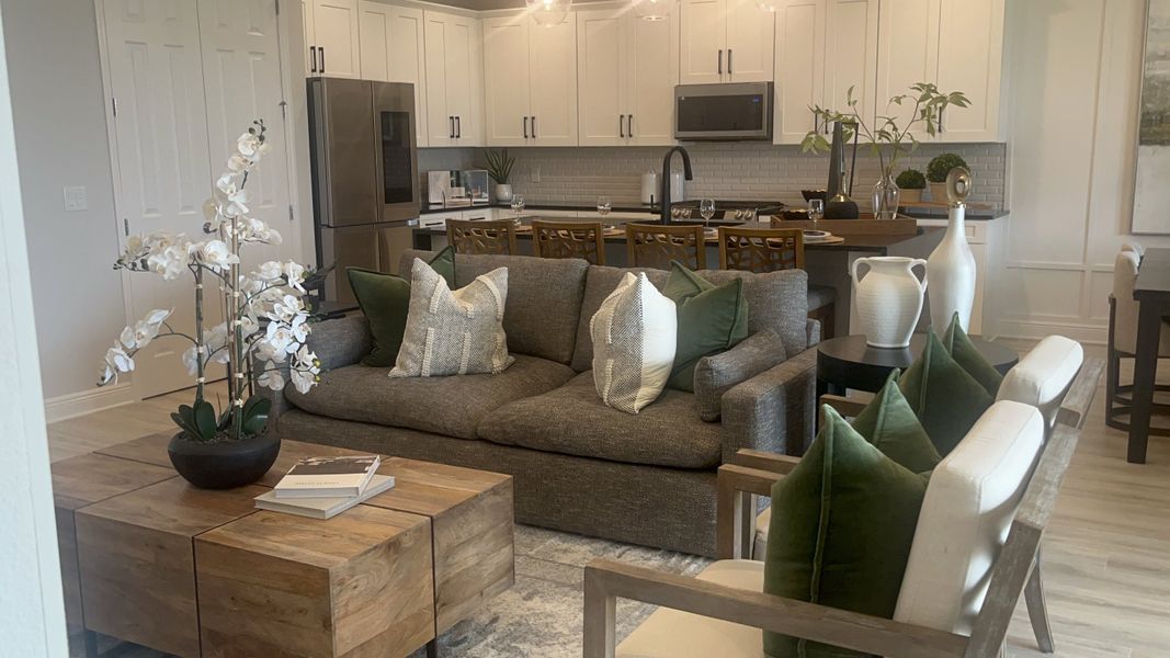 A cozy living area with a modern kitchen, plush gray sofa, and elegant wooden accents complemented by green pillows and plants.