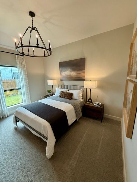 A cozy bedroom with a modern chandelier, plush carpet, and elegant decor, featuring a large window for natural light.