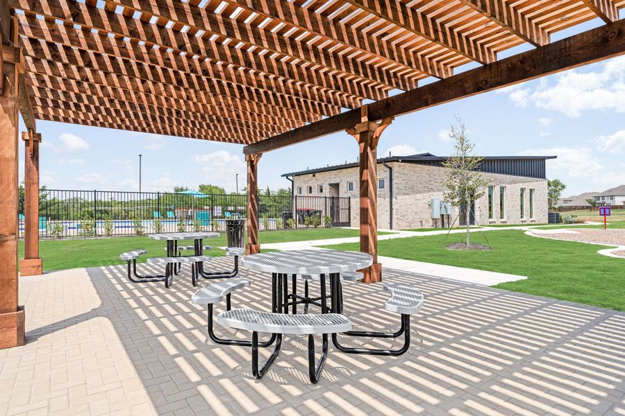 Outdoor pergola with metal picnic tables on a paver patio, community pool and stone building in the background.