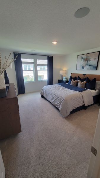A cozy bedroom featuring a plush bed, soft carpet, and stylish decor with ample natural light filtering through large windows.